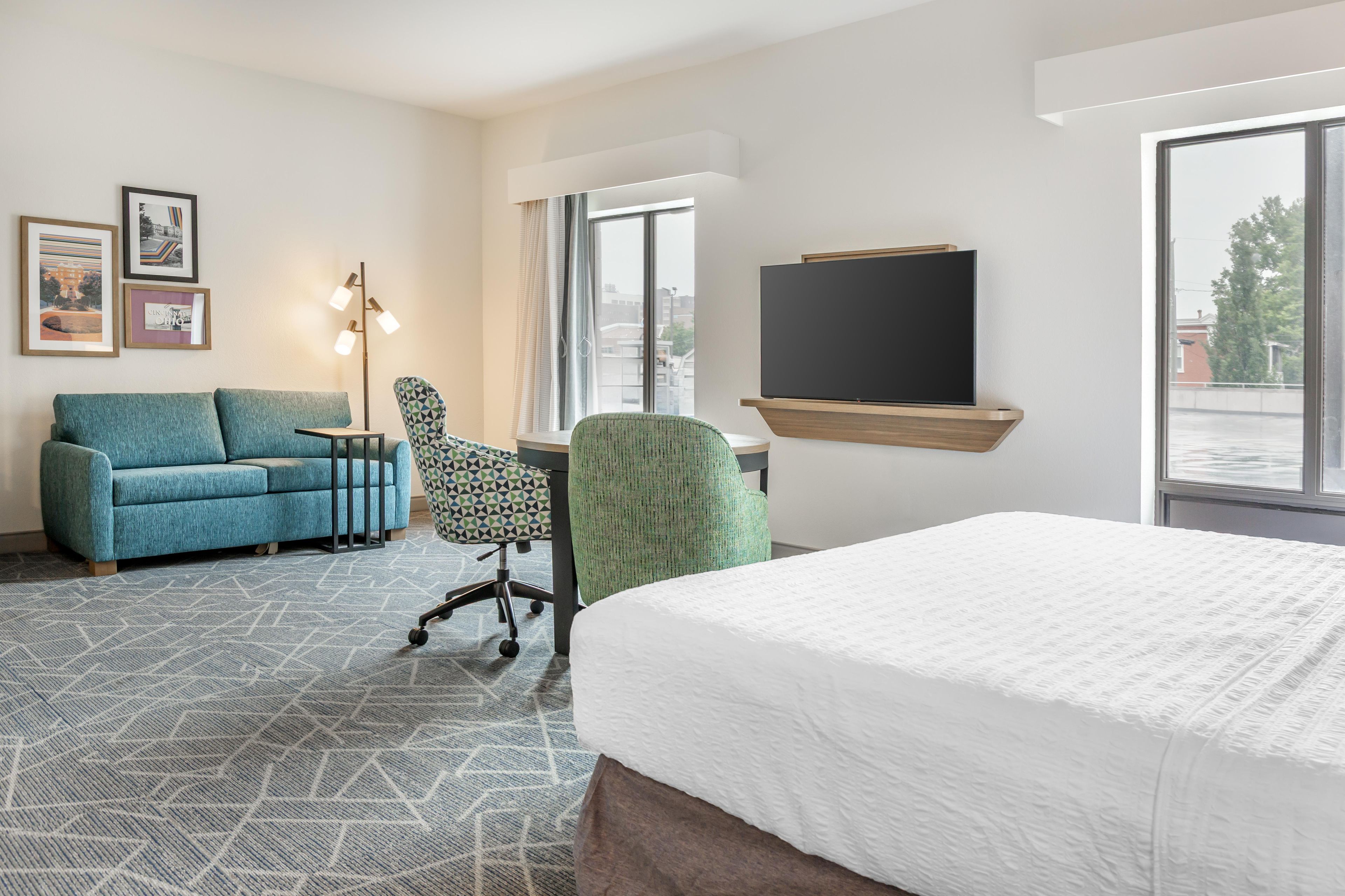 A hotel room with a bed, a sofa, a desk, and a chair. There is a flat-screen TV mounted on the wall and a window with a view of the outside.