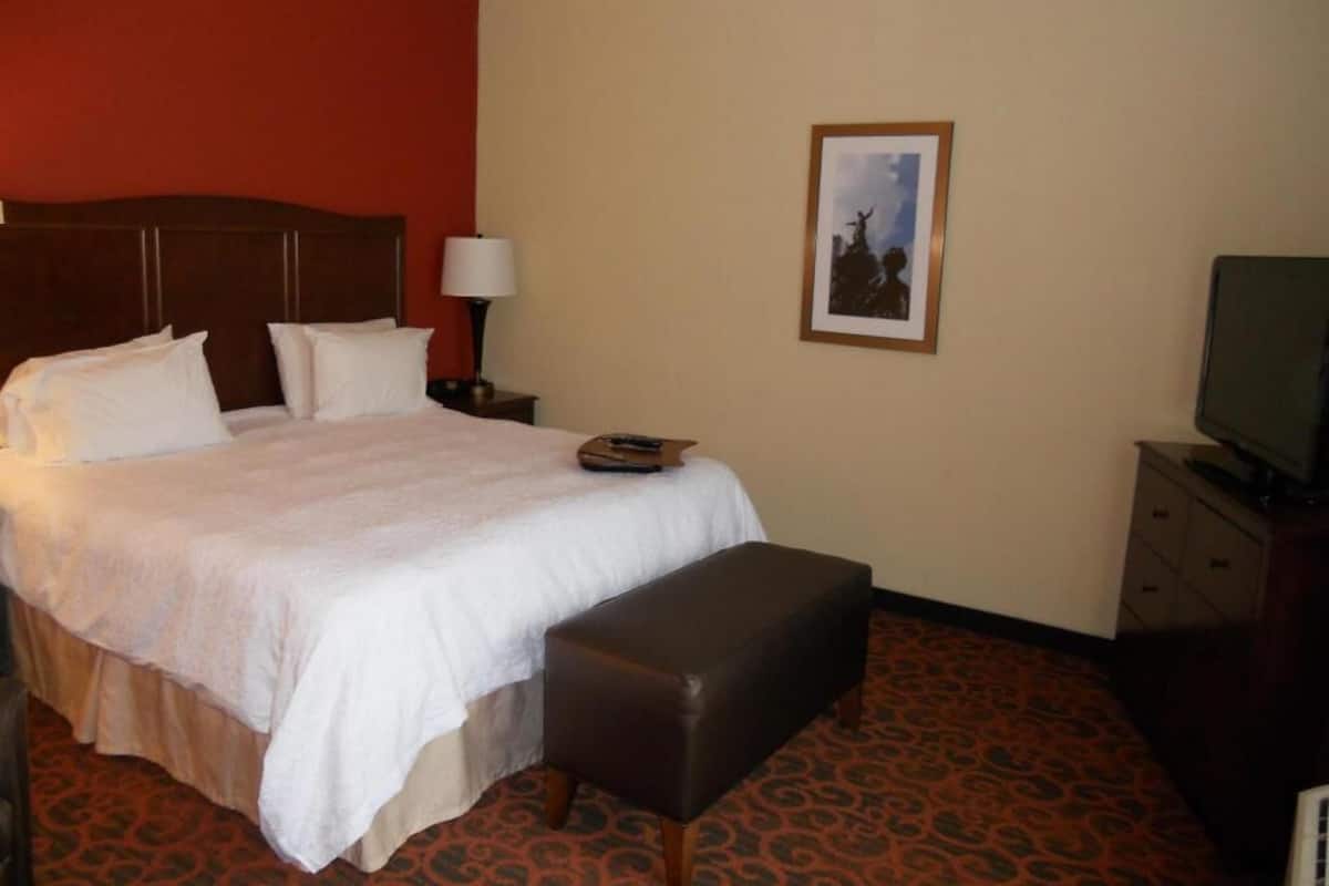 A hotel room with a bed, a bench, a nightstand, a lamp, a television, and a framed picture on the wall.
