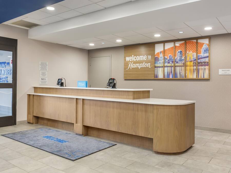 Zona de recepción de un hotel con un mostrador de madera, un teléfono digital y un cartel que dice 'Welcome to Hampton'.