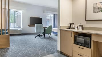Una habitación de hotel moderna con kitchenette, escritorio, silla y un ventanal grande.