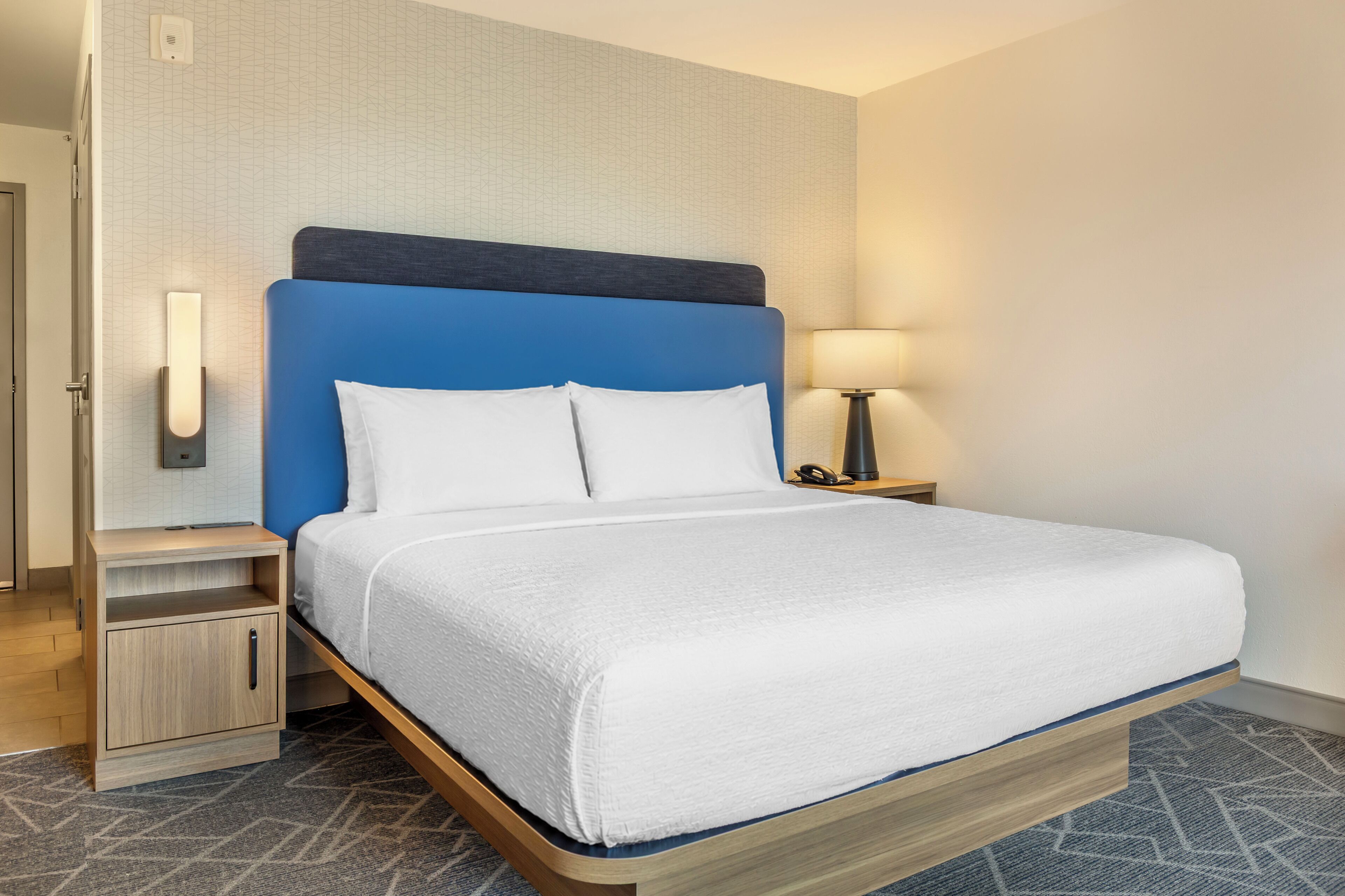 Habitación de hotel con cama, mesita de noche, lámpara y teléfono instalado en la pared.