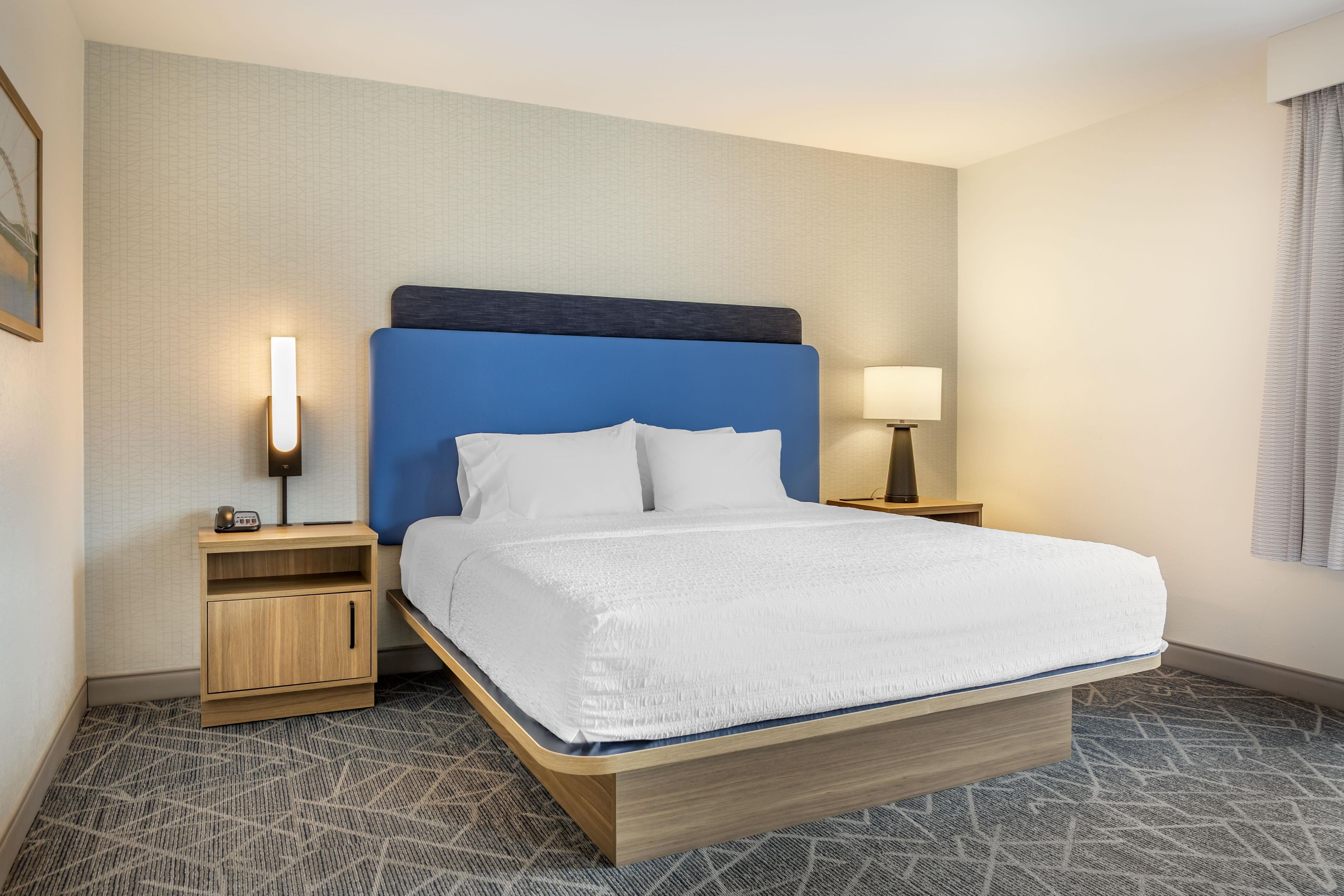 A hotel room with a bed, a nightstand, a lamp, and a wall-mounted light.