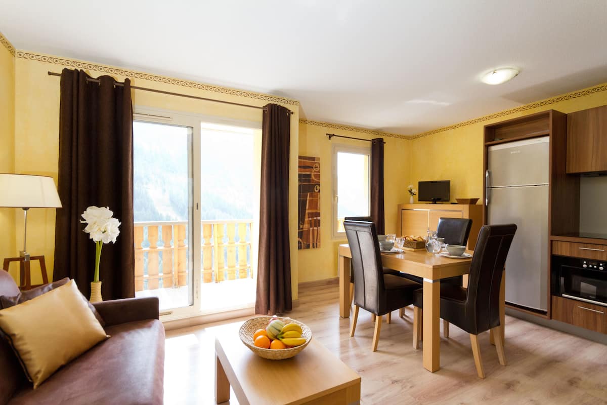 Ein Wohnzimmer mit einer Couch, einem Esstisch mit Stühlen, einer Küchenzeile inklusive Kühlschrank und einem Balkon mit Vorhängen.