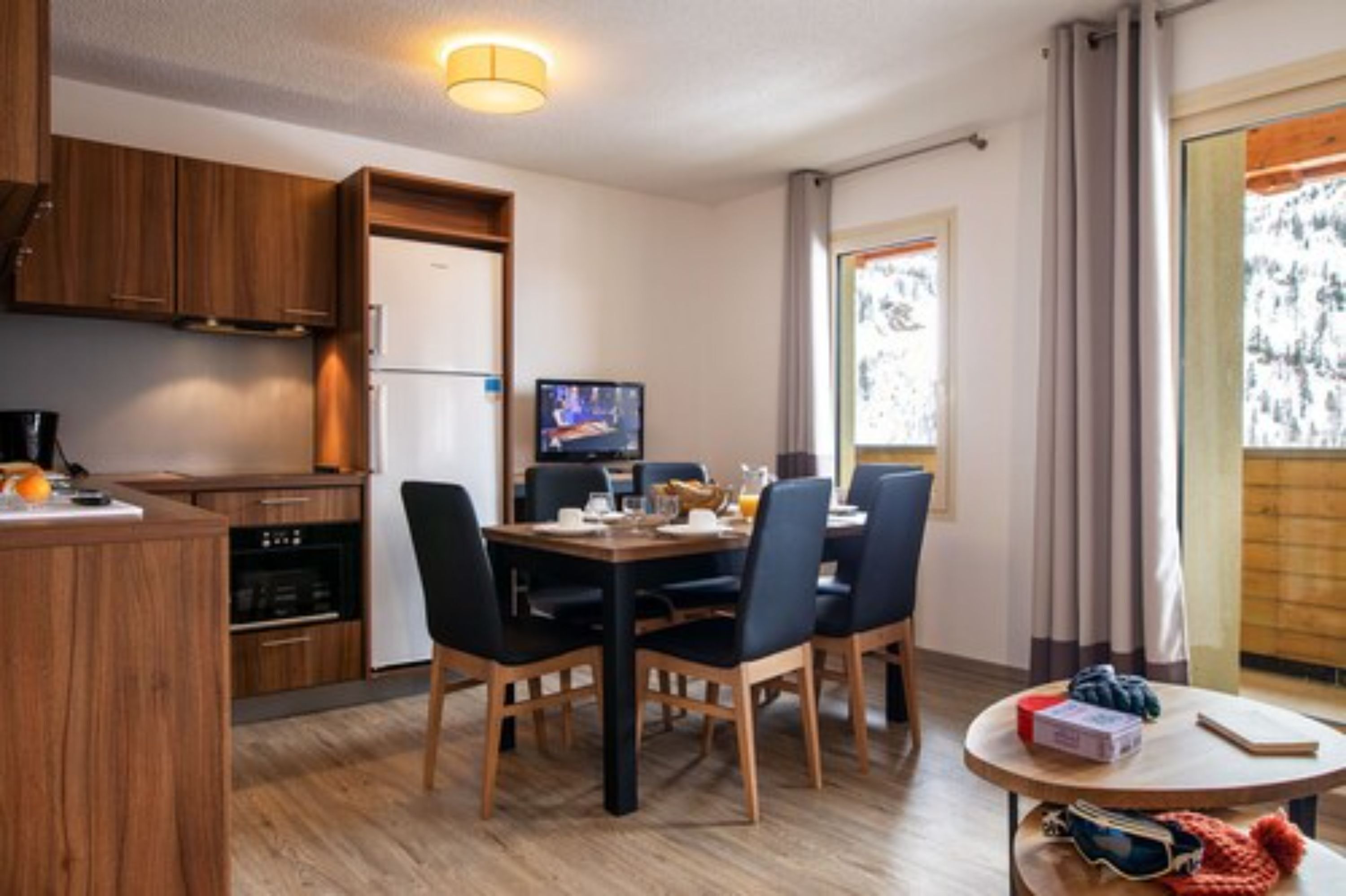 Une cuisine moderne avec des meubles en bois, un coin repas avec une table et des chaises, un téléviseur et une fenêtre avec des rideaux.