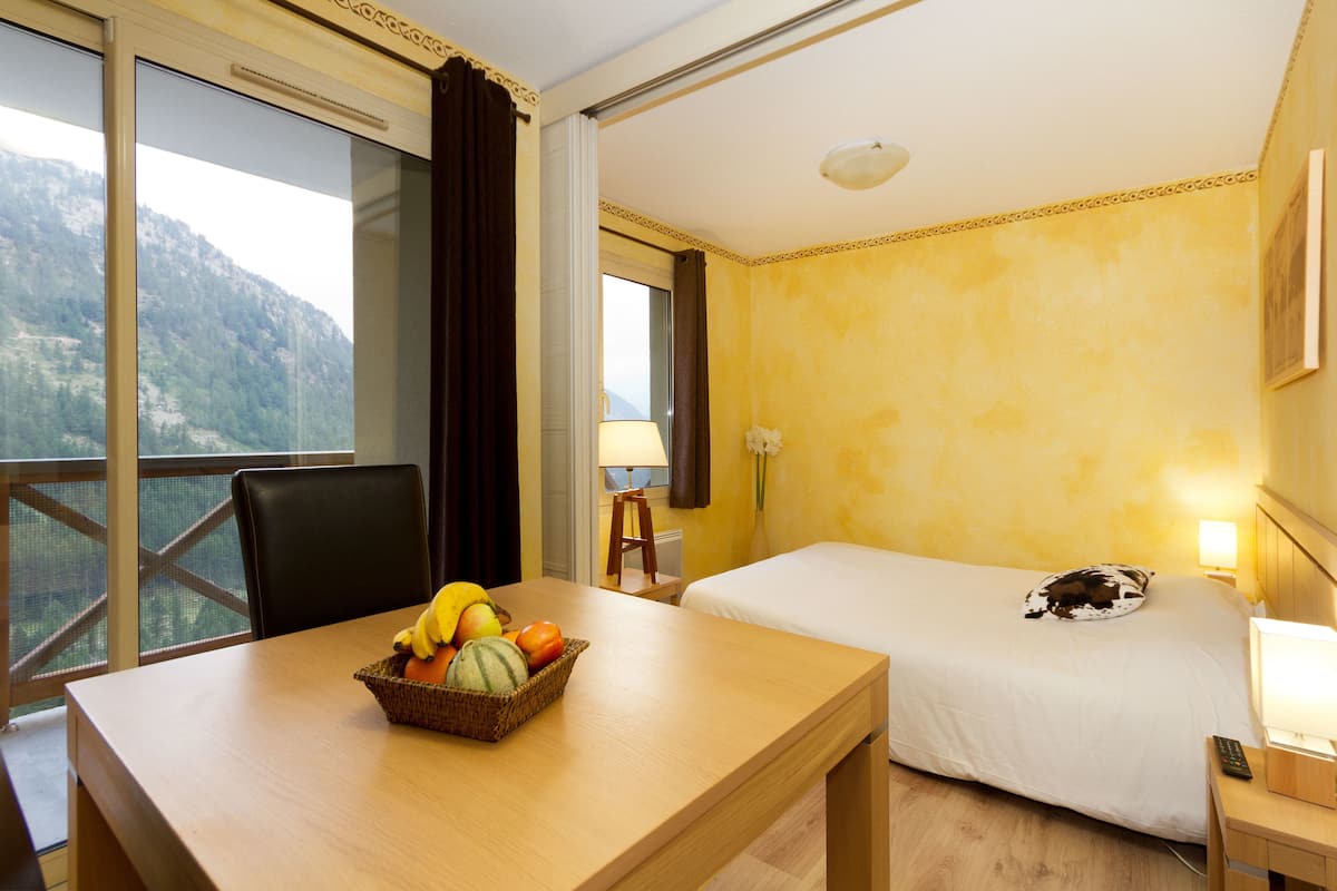 Ein Hotelzimmer mit einem Bett, einem Schreibtisch mit Obstkorb, einem Sessel, einer Lampe und Blick auf Berge.