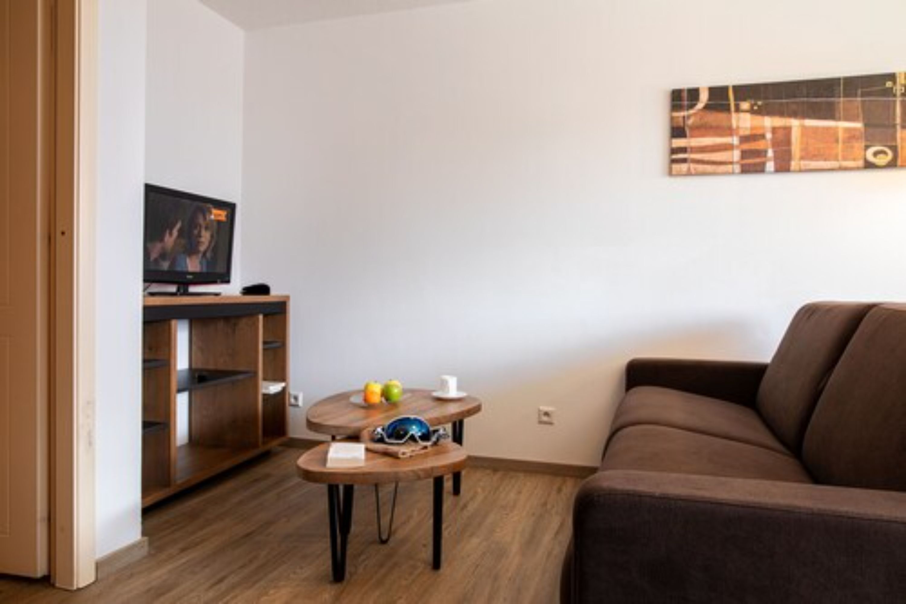 Una sala de estar moderna con un sofá marrón, un mueble de madera para entretenimiento, un televisor y una pequeña mesa redonda con una taza y gafas de sol.