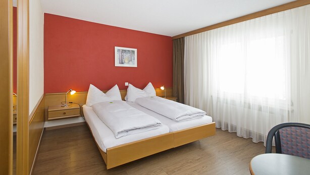 A hotel room with a wooden bed, two bedside tables with lamps, a red accent wall, a window with sheer curtains, and a chair. - Hotel Sportcenter Fünf Dörfer (Trimmis)