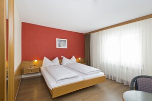 A hotel room with a wooden bed, two bedside tables with lamps, a red accent wall, a window with sheer curtains, and a chair. - Hotel Sportcenter Fünf Dörfer (Trimmis)