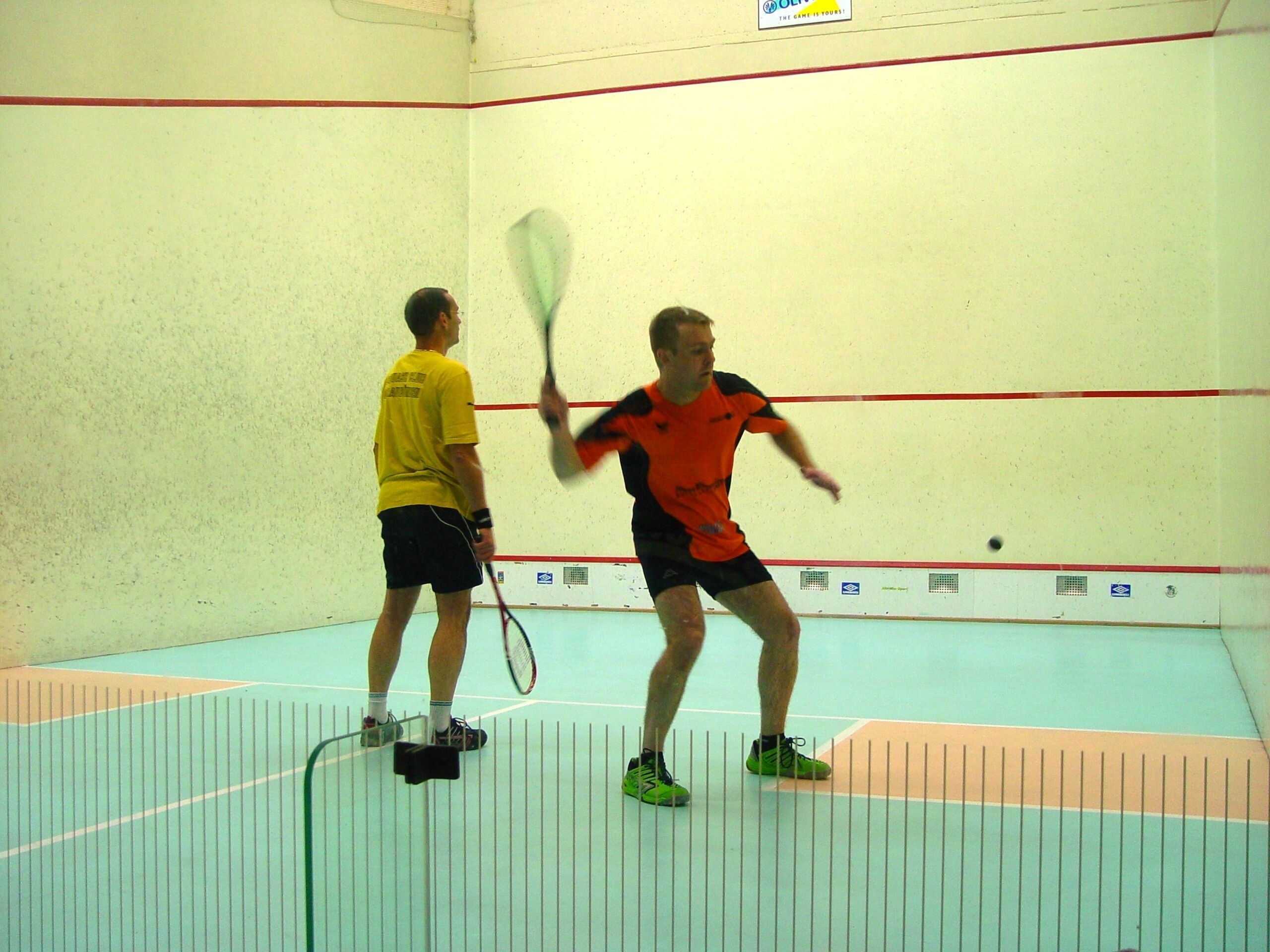Deux personnes jouent au squash dans un court intérieur, avec un mur visible et une porte.