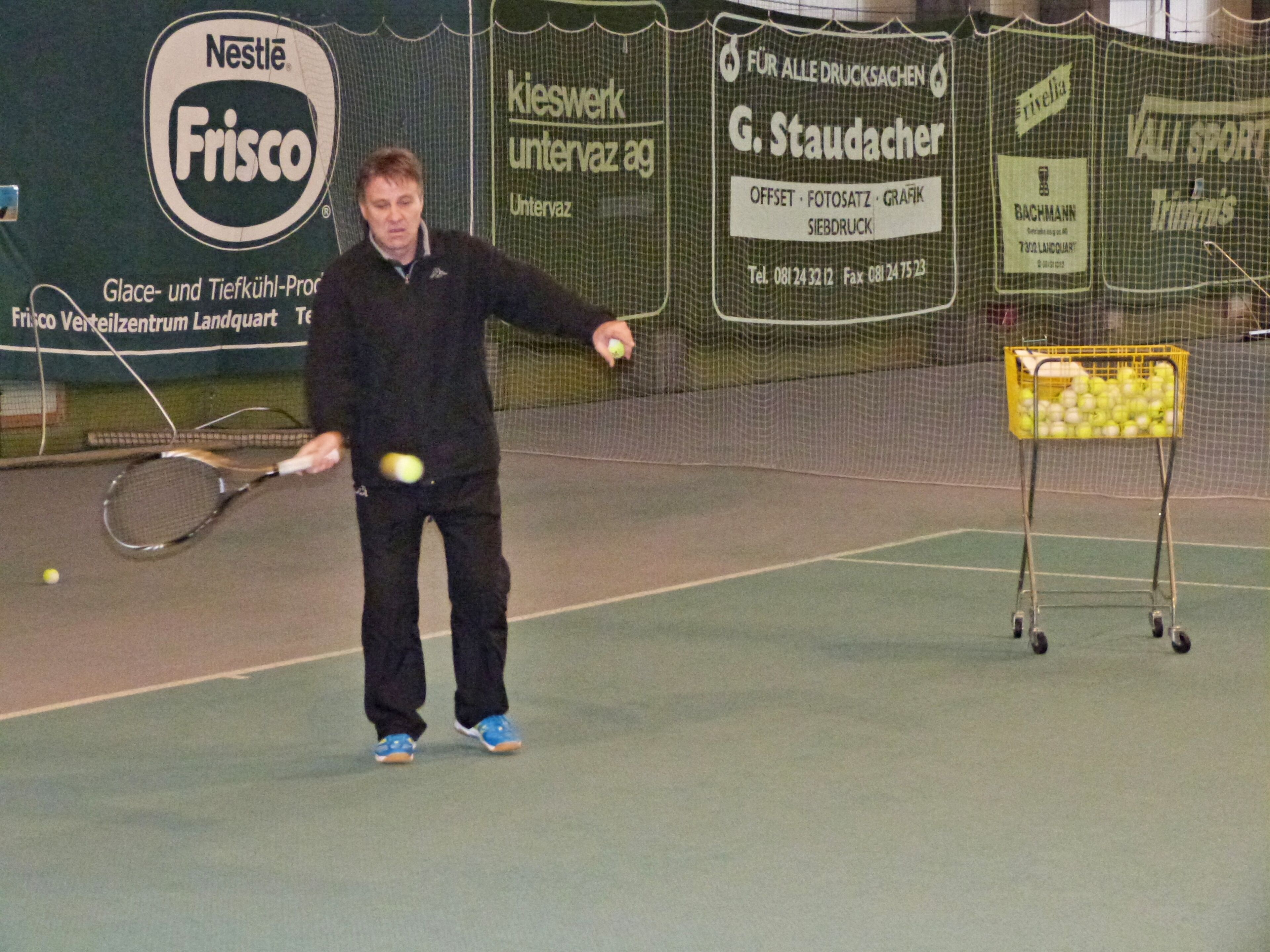 A tennis court with a net, a tennis player, and a basket of tennis balls.
