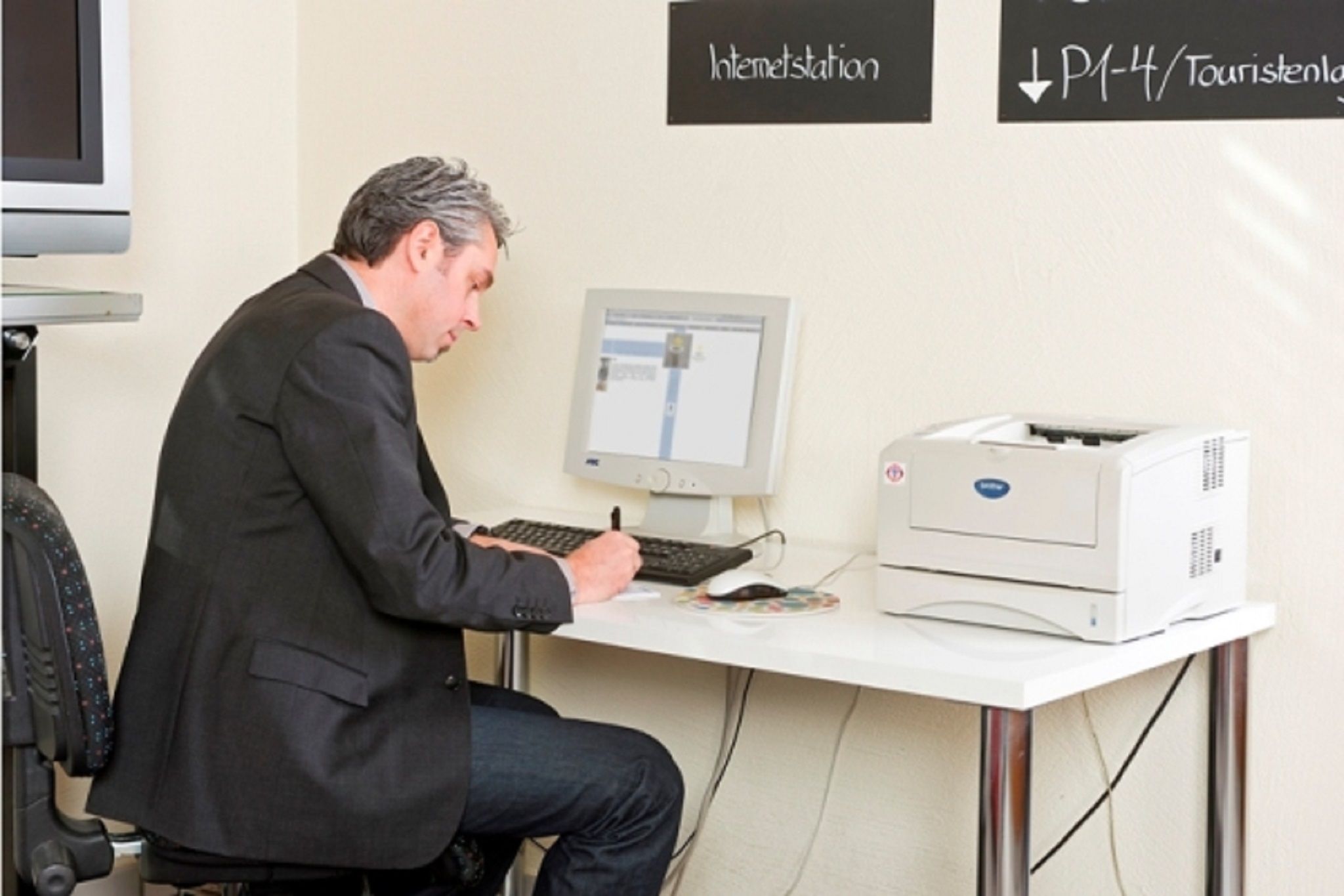 A man sitting at a desk with a computer, a printer, and a monitor. There are signs on the wall indicating "Internetstation" and "P1-4/Touristenland".
