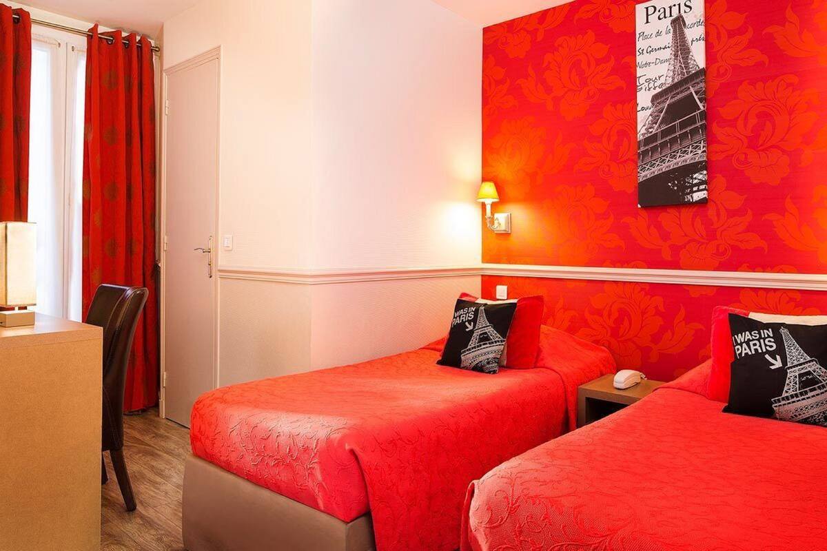 A hotel room with two beds, a desk, and a chair. There is a wall-mounted lamp and a framed picture of the Eiffel Tower.