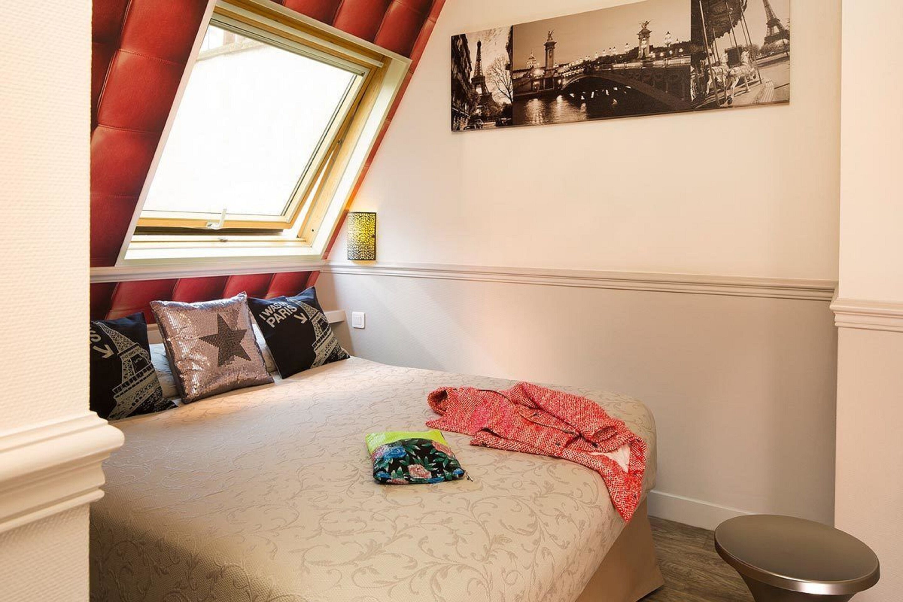 A bedroom with a bed, a window, a red curtain, a picture of Paris, and a small stool.