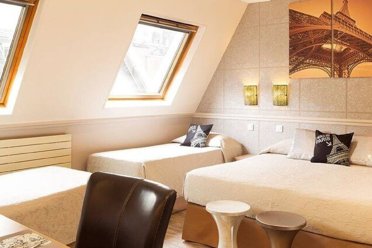A hotel room with two beds, a desk, a chair, and a small table. There are windows with views of the outside.