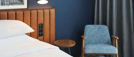 Quarto de hotel com uma cama, uma luminária fixada na parede, uma mesa redonda pequena, uma poltrona azul estofada e um quadro na parede.