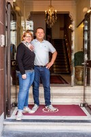 A man and woman standing at the entrance of a hotel with a red carpet, a chandelier, and a staircase in the background.
