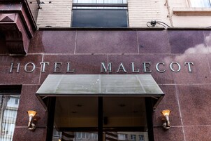 Η είσοδος του ξενοδοχείου Malecot, με μια μεγάλη, εμφανή πινακίδα και δύο φωτιστικά σώματα τοποθετημένα στους τοίχους.