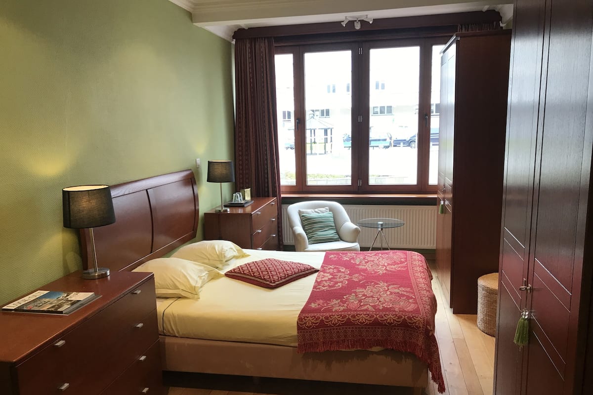 Ein Hotelzimmer mit Bett, Sessel, kleinem Tisch, Lampe und Fenster mit Außenblick.