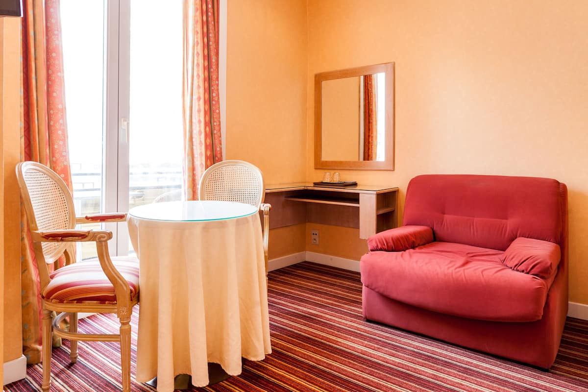 Ein Hotelzimmer mit Streifenmuster-Teppich, einem runden Tisch mit zwei Stühlen, einem roten Sessel, einem Schreibtisch mit Stuhl und einem großen Spiegel an der Wand.