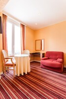 A hotel room with a striped carpet, a round table with two chairs, a red armchair, a desk with a chair, and a large mirror on the wall.