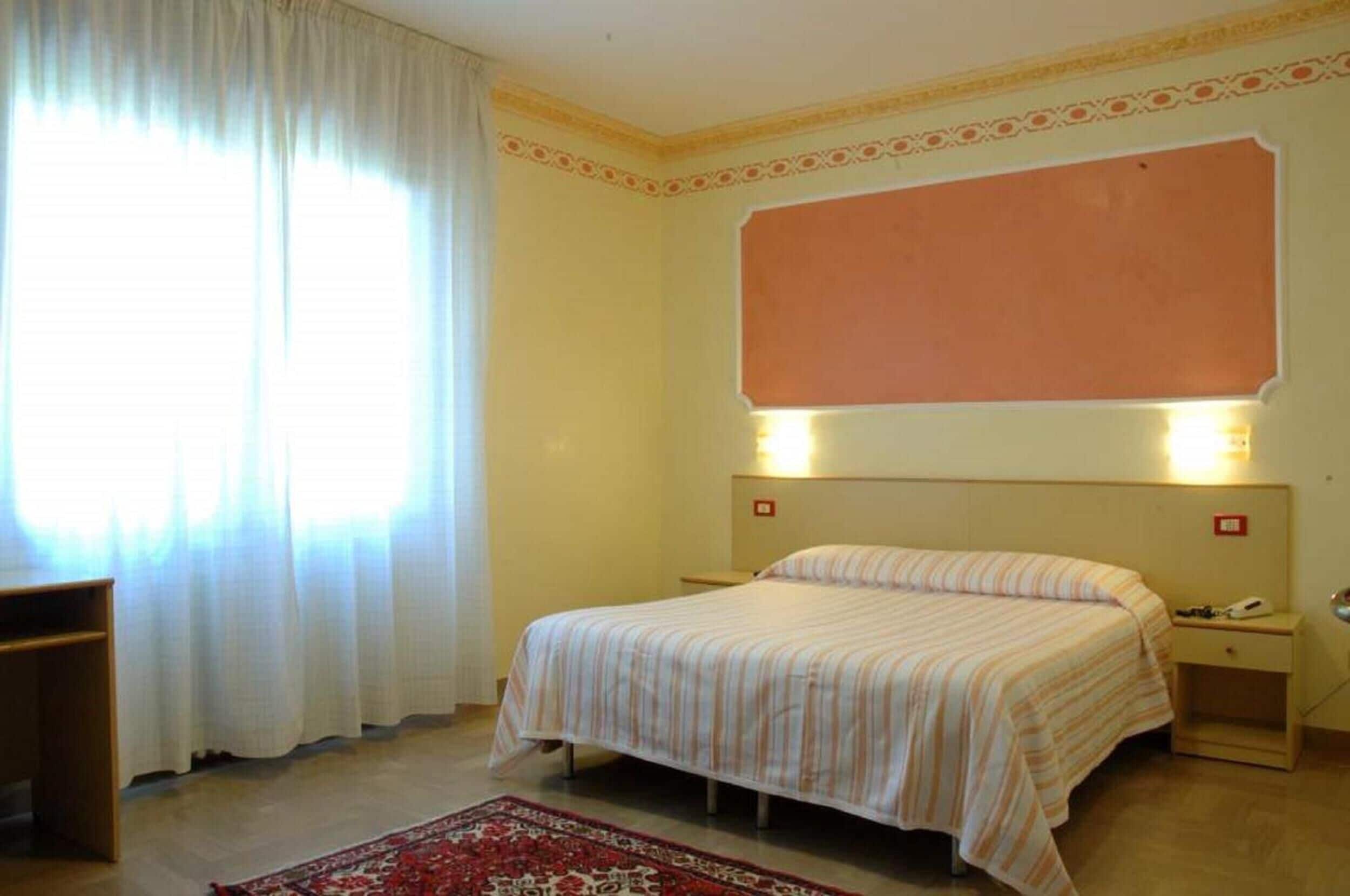 Habitación de hotel con cama, mesitas de noche, alfombra roja estampada y un ventanal con cortinas blancas.
