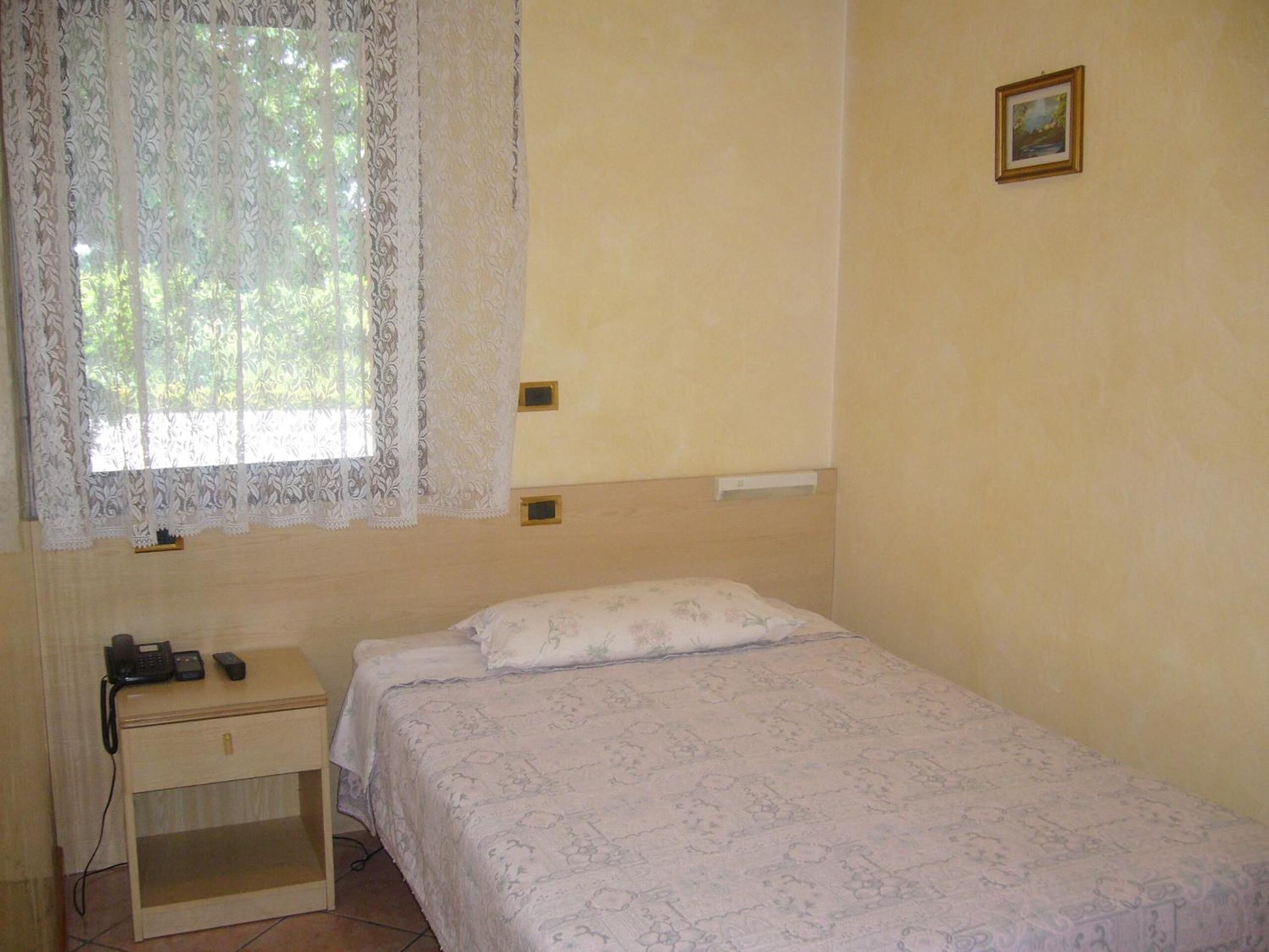 Un dormitorio pequeño y sencillo con una cama individual, una mesita de noche, un teléfono y una ventana con cortinas de encaje.