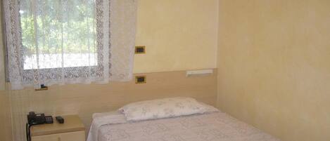 Un dormitorio pequeño y sencillo con una cama individual, una mesita de noche, un teléfono y una ventana con cortinas de encaje.