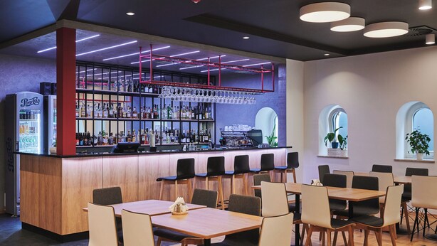 A modern bar area with a wooden counter, shelves of bottles, and a refrigerated display. Tables and chairs are arranged for dining.