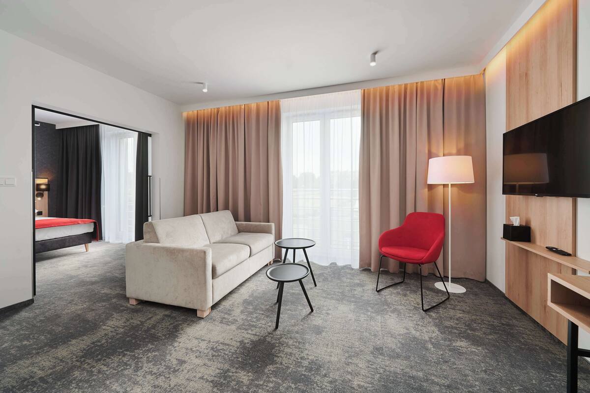 A modern hotel room with a sofa, a red chair, a small table, a TV, and a large window with curtains.