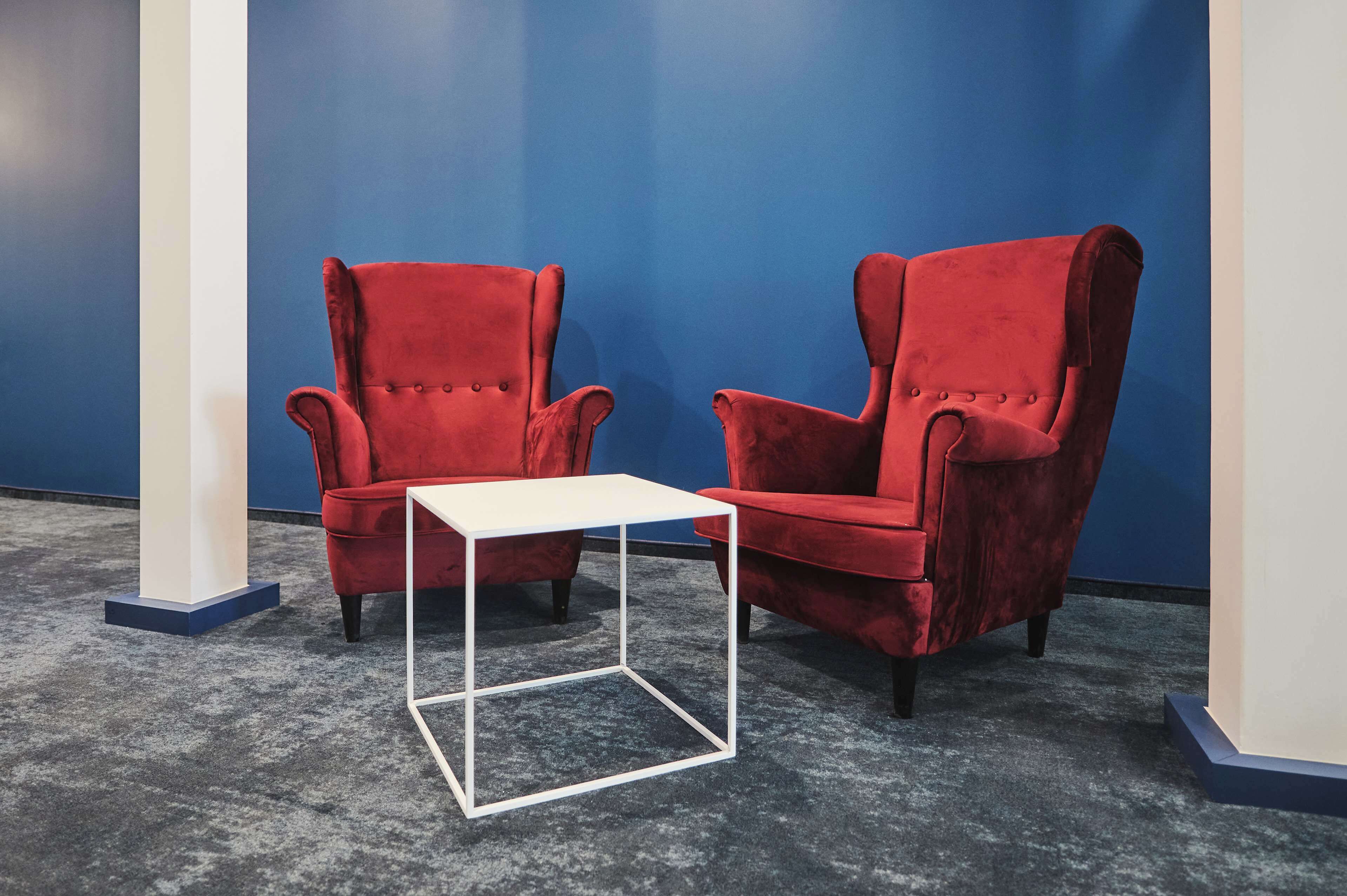Two red armchairs and a white table in a room with blue walls and a grey carpet.