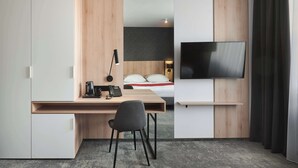 A compact hotel room with a wooden desk, a chair, a bed with white bedding, a flat-screen TV, and a large window with curtains.