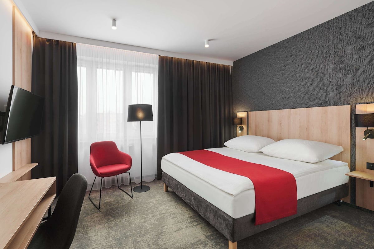 A modern hotel room with a large bed, a desk, a chair, and a TV.