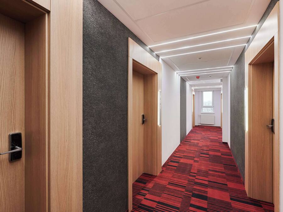 Un pasillo moderno de hotel con puertas de madera, una alfombra roja estampada e iluminación empotrada.