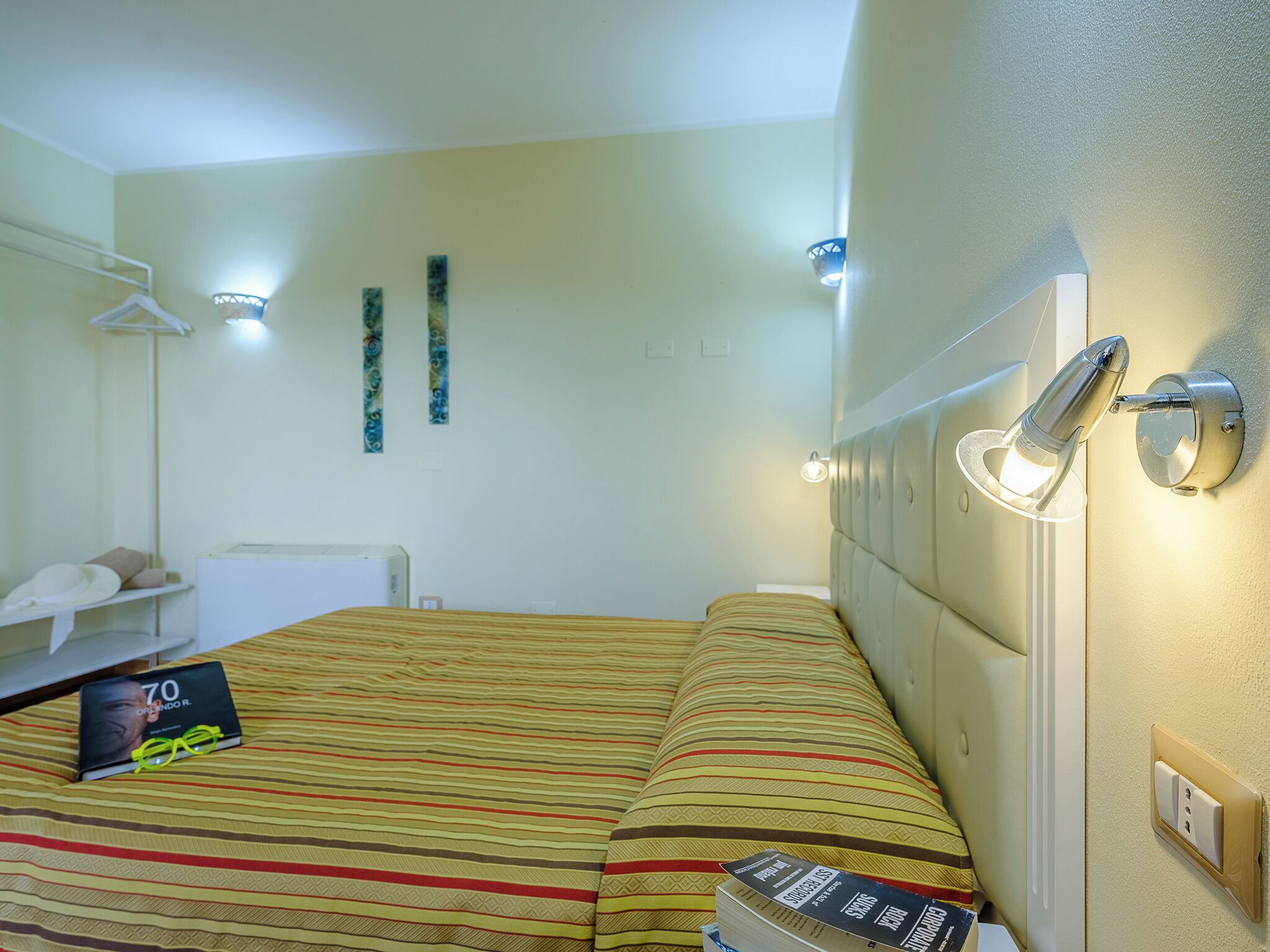 Quarto de hotel com cama, mesa de cabeceira, abajur e luminária na parede.