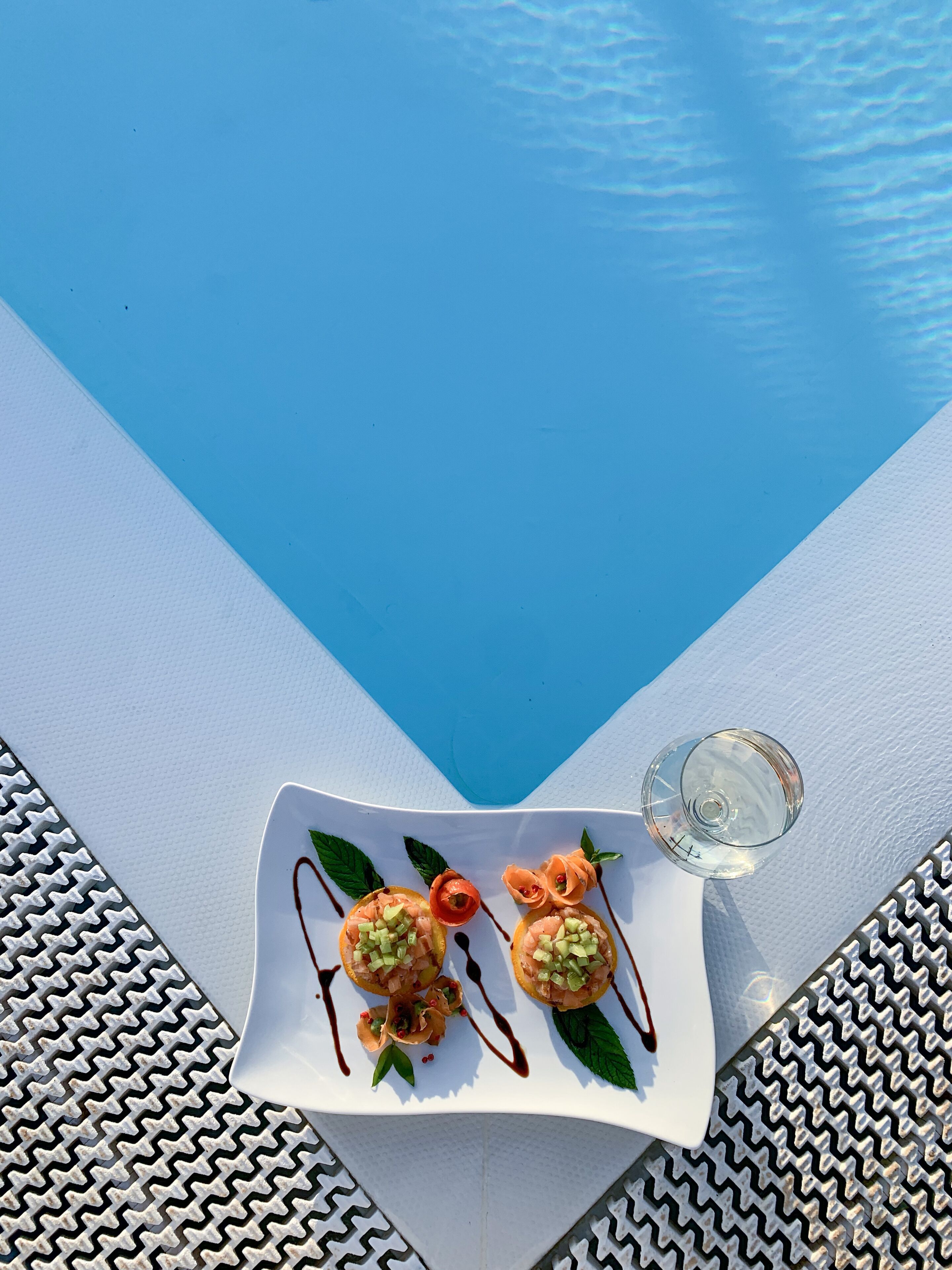 A white plate with food and a glass of water on a white and blue striped surface near a pool.