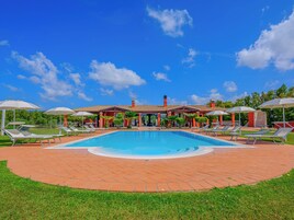 Una piscina con lettini e ombrelloni, circondata da un patio in mattoni e vegetazione.