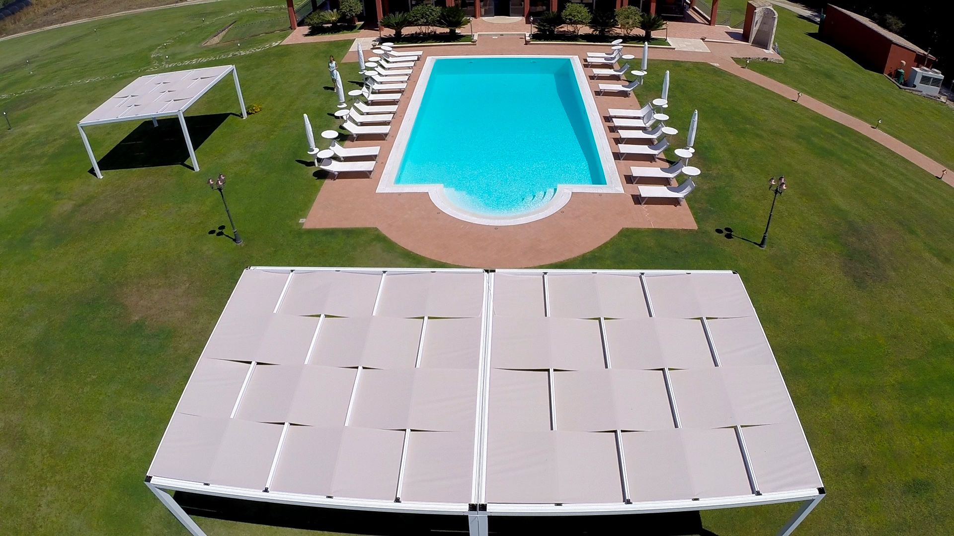 Vue aérienne d’une piscine entourée de chaises longues et de parasols, elle-même bordée d’une pelouse bien entretenue et d’un espace de détente abrité.