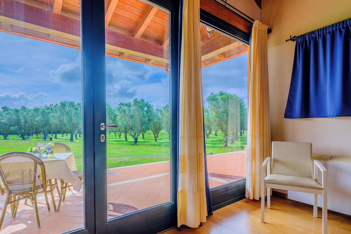 Una stanza con un'ampia porta-finestra che dà su un patio esterno con tavolo e sedie, vista su una zona verde alberata e una tenda blu alla destra.