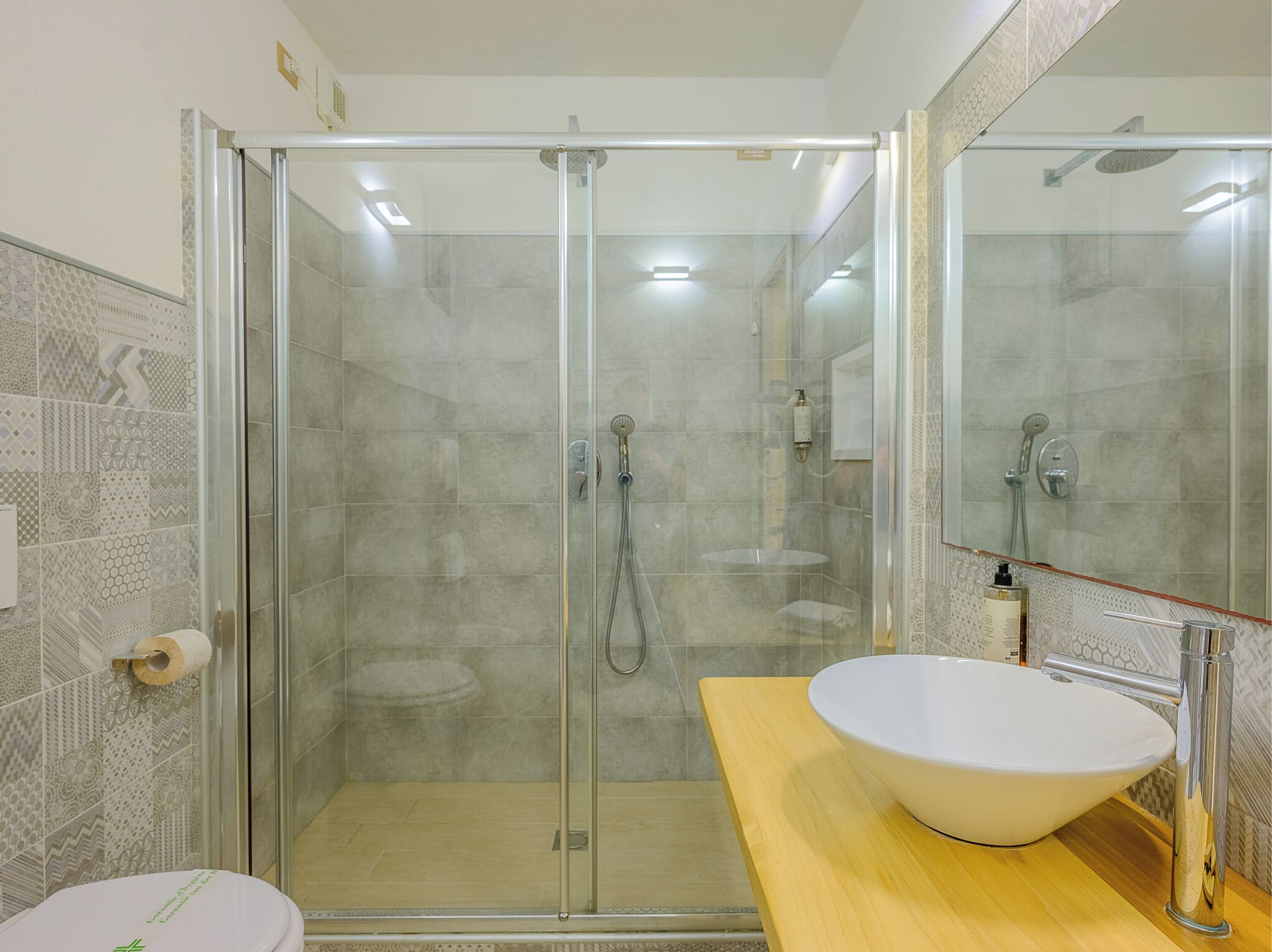 Un baño moderno con cabina de ducha de vidrio, lavamanos blanco y baldosas estampadas.