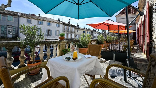 An outdoor terrace with a table set for two, covered by a large blue umbrella, surrounded by wicker chairs, and overlooking a street with buildings. - Hôtel des Artistes (Tarascon)