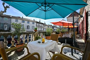 An outdoor terrace with a table set for two, covered by a large blue umbrella, surrounded by wicker chairs, and overlooking a street with buildings. - Hôtel des Artistes (Tarascon)