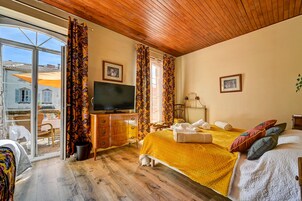 Un dormitorio con techo de madera, una cama con manta amarilla, un televisor sobre un aparador de madera y vista a un patio a través de un vidrio.