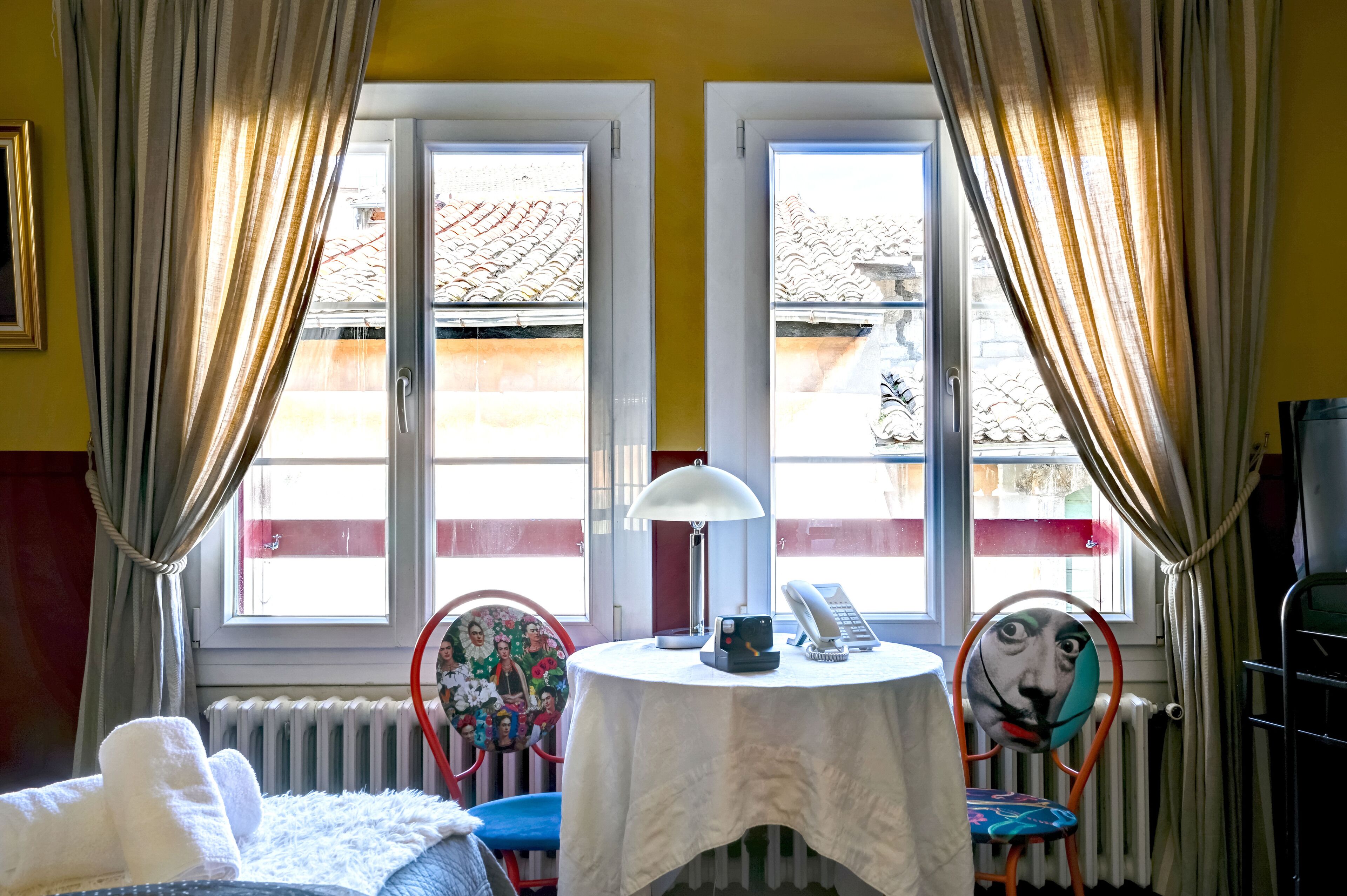 Una habitación con cama, una mesa con lámpara, sillas y vistas a edificios a través de ventanas.