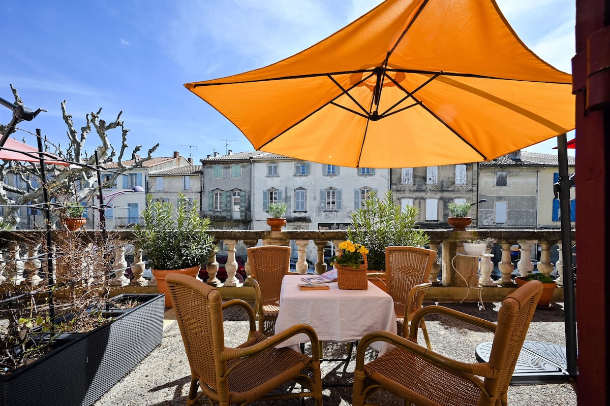 An outdoor seating area with wicker chairs, a table with a white tablecloth, and an orange umbrella providing shade.