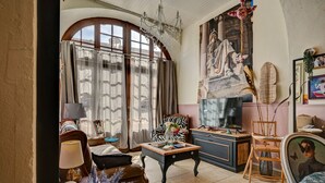 A spacious living room with a large arched window, a chandelier, a television, and a painting on the wall. - Hôtel des Artistes (Tarascon)