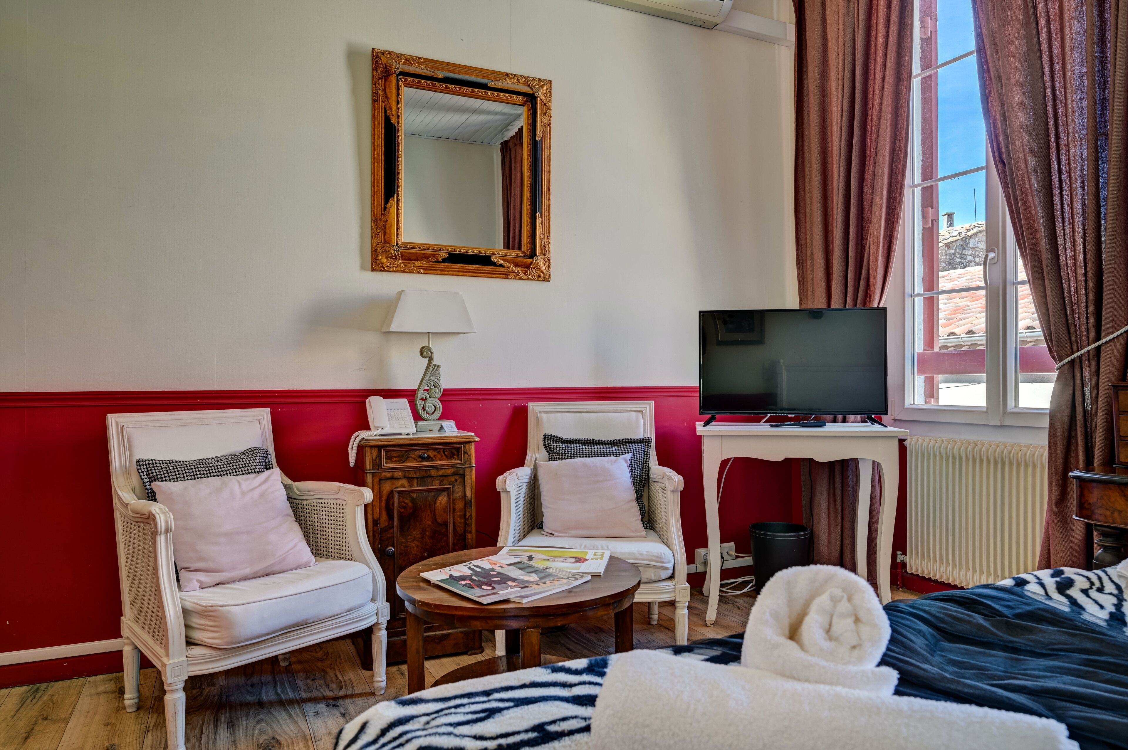 A hotel room with two white wicker chairs, a small round table with a lamp, a television on a stand, and a bed with a zebra-patterned blanket.