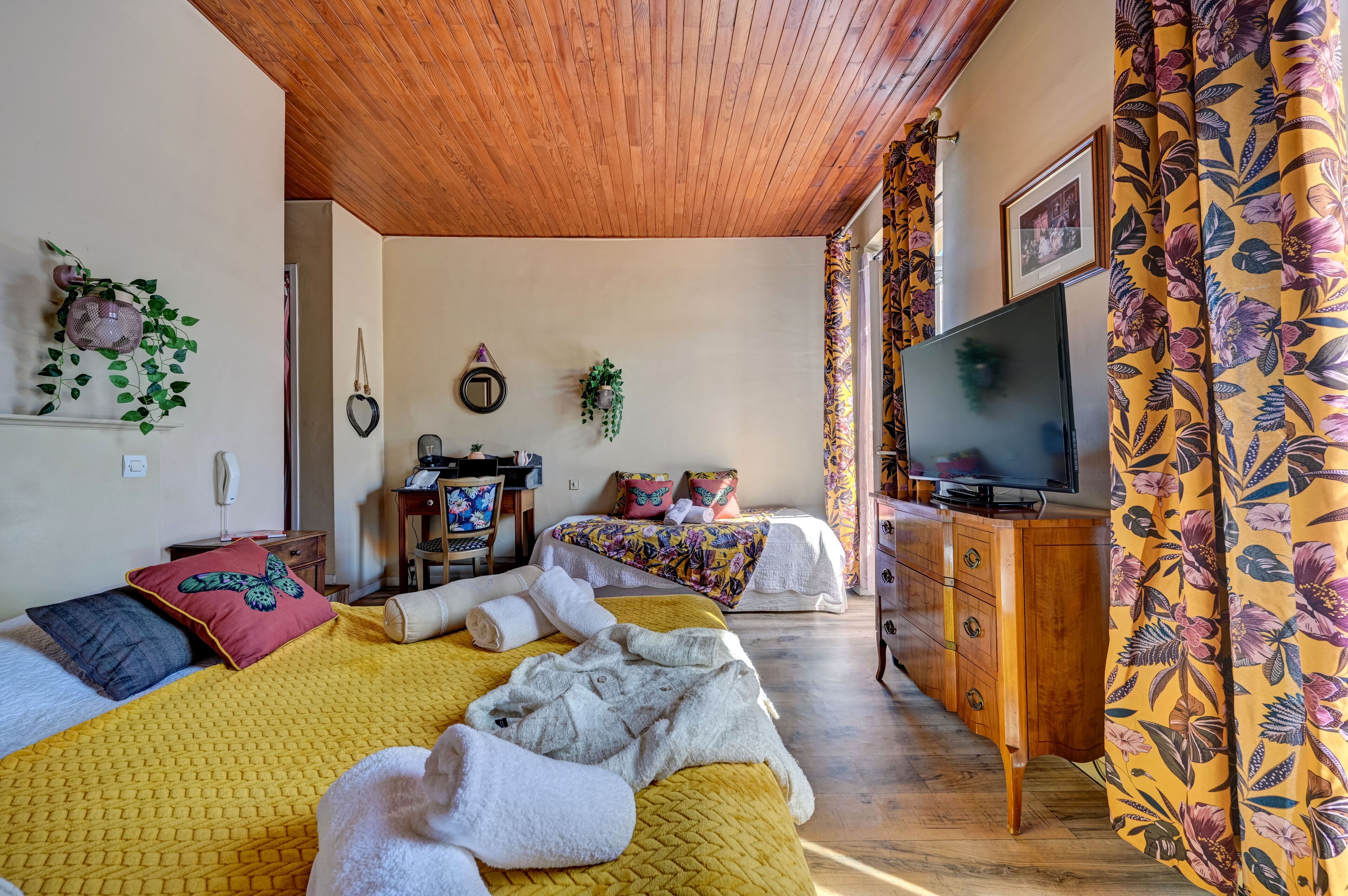 A room with two beds, a wooden ceiling, a TV, and a floral curtain.