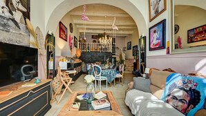 A spacious living room with a large TV, a wooden coffee table, and a sofa with a colorful throw. There is a dining area with a table and chairs, and a staircase leading to an upper level. - Hôtel des Artistes (Tarascon)