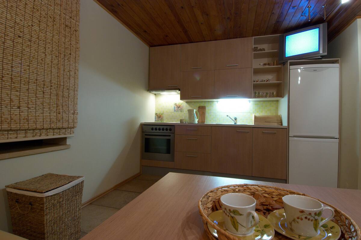 Una cocina con armarios de madera, una estufa, un refrigerador, un televisor y una mesa con una bandeja de tazas y platillos.
