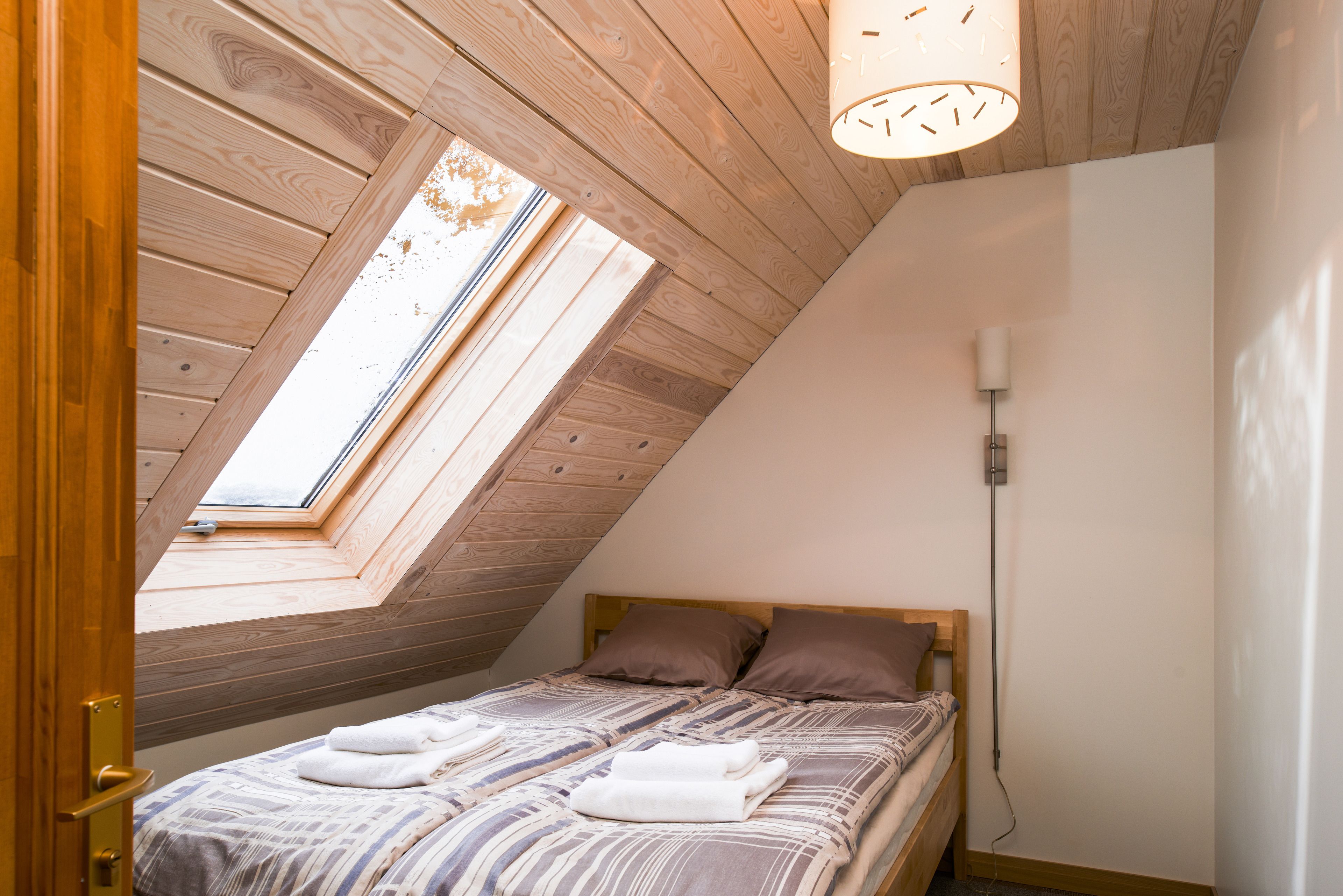 A bedroom with a sloped ceiling, a skylight, a bed with pillows, and a bedside lamp.
